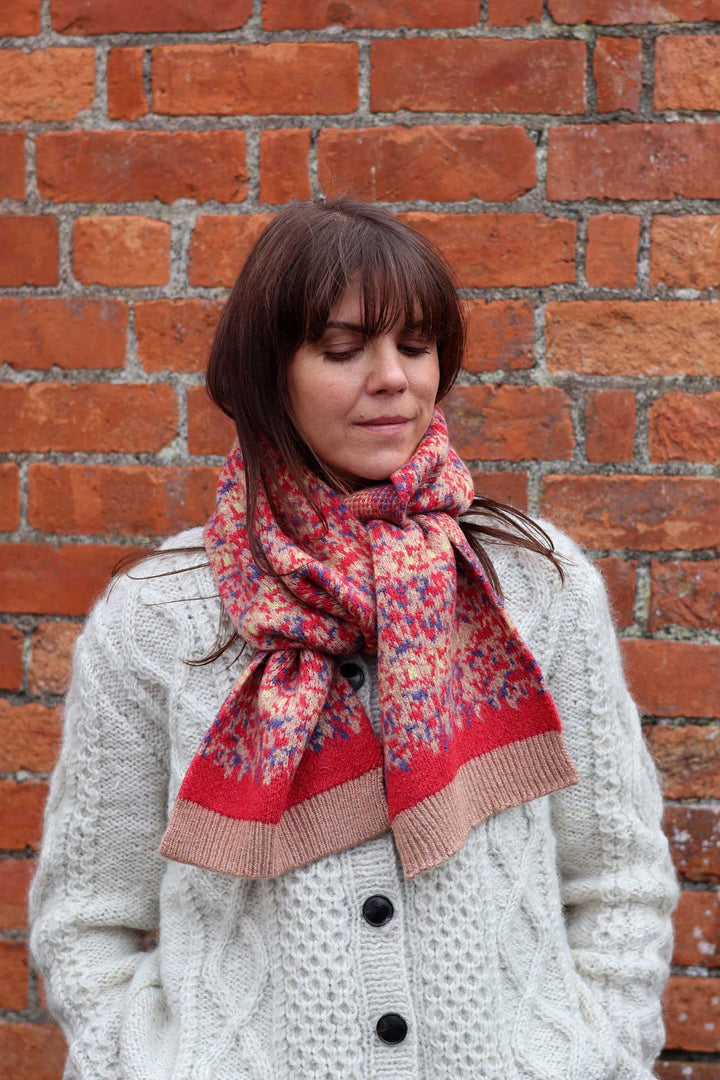 A woman wearing a cream cable-knit cardigan and a red, pink, and blue patterned knit scarf, standing in front of a red brick wall.
