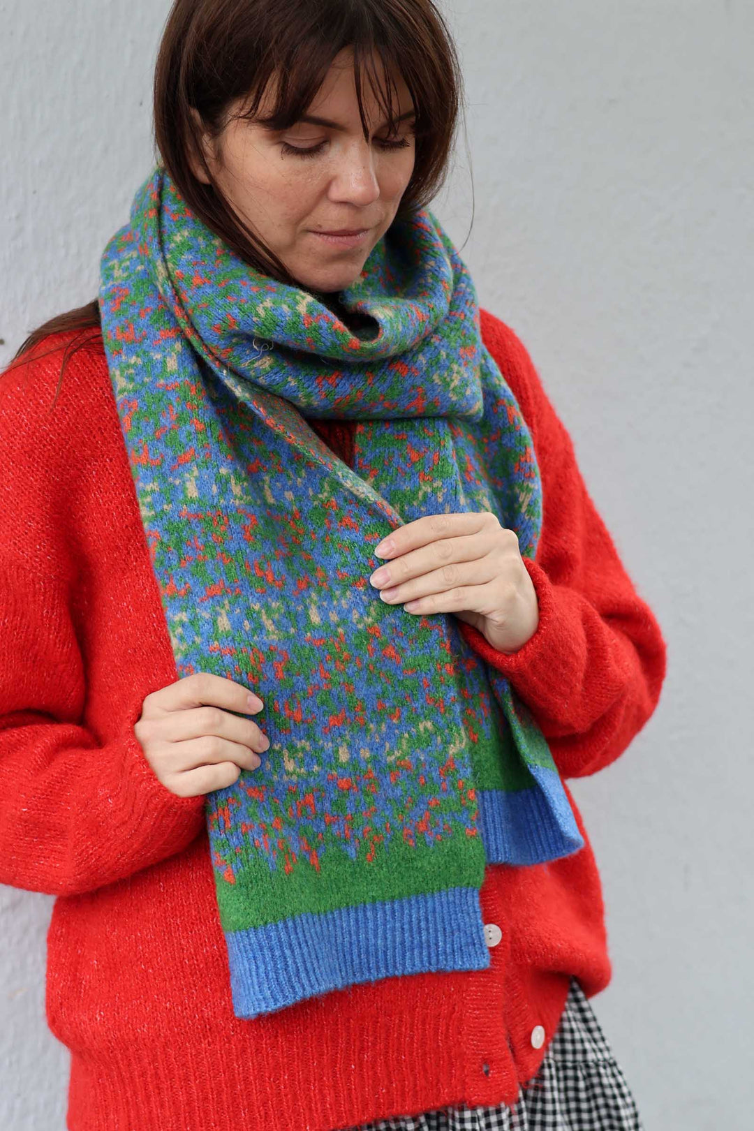 A woman wearing a bright red cardigan and a multicolored knitted scarf with blue ribbed ends, standing against a plain light gray wall.