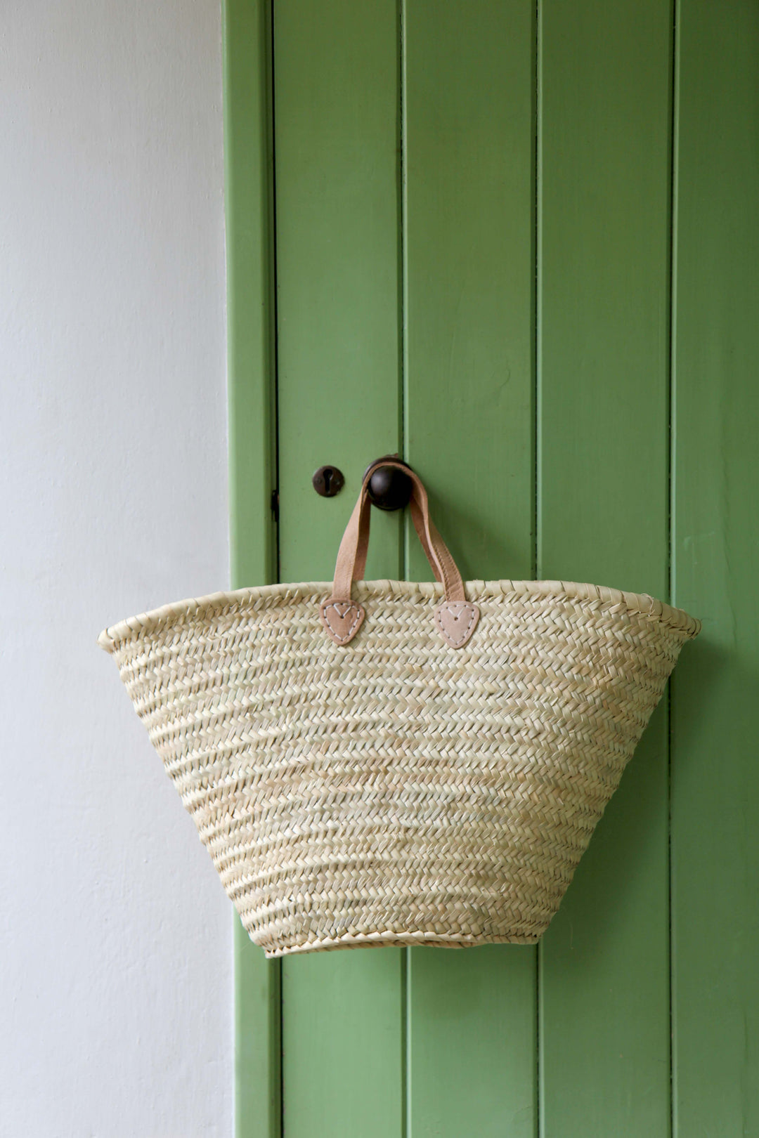 Woven basket with leather handles hanging on a green door.