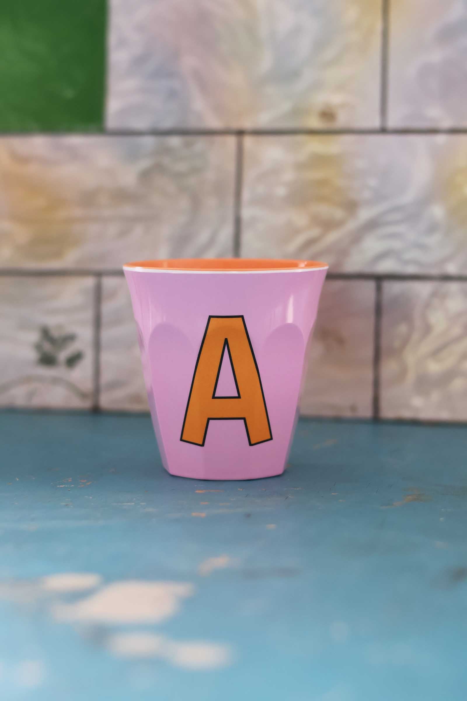 Pink cup with an orange letter 'A' on a blue surface with a tiled wall background
