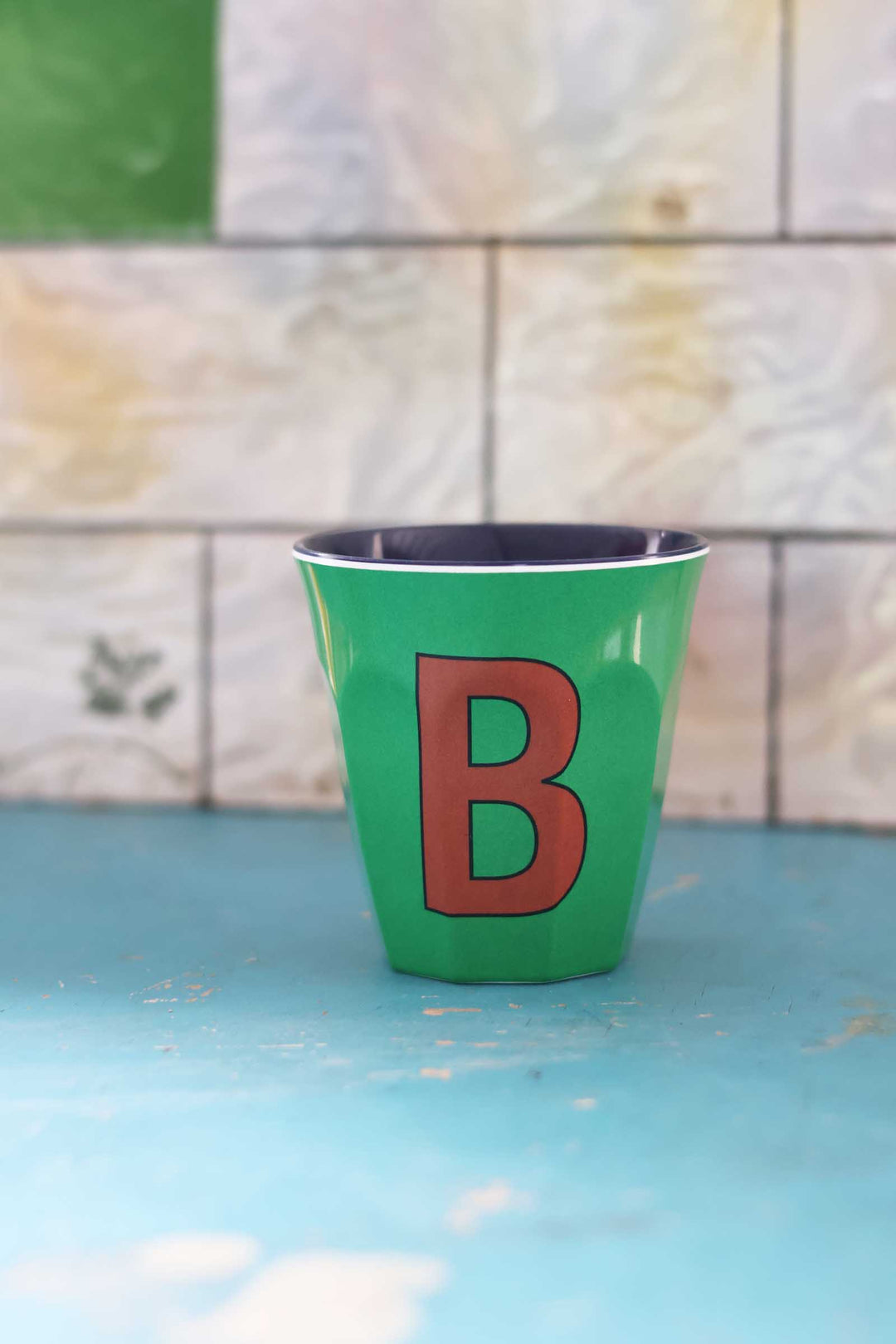 Green cup with a red letter 'B' on a blue surface with a tiled wall background