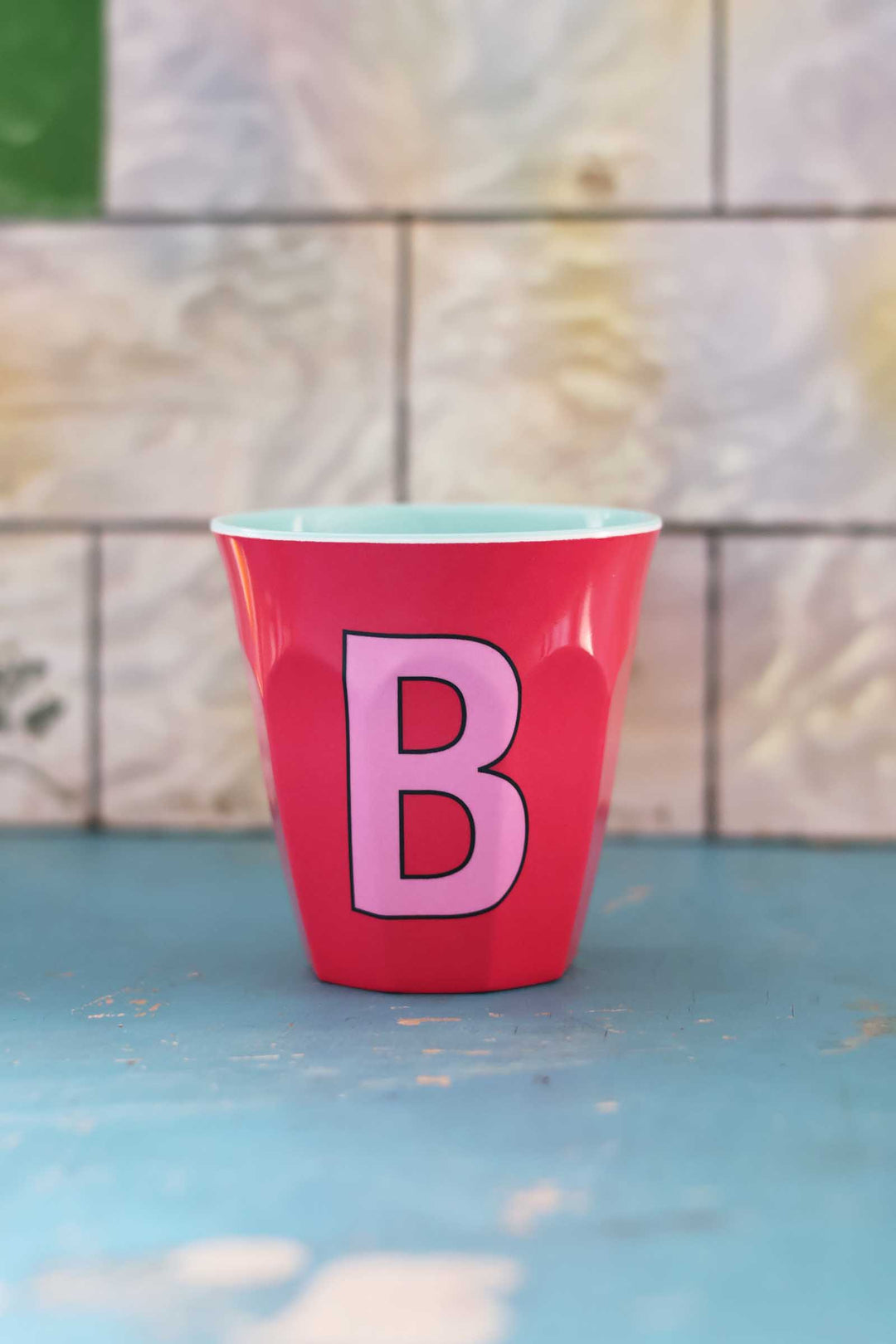 Red cup with a pink letter 'B' on a blue surface and tiled wall background