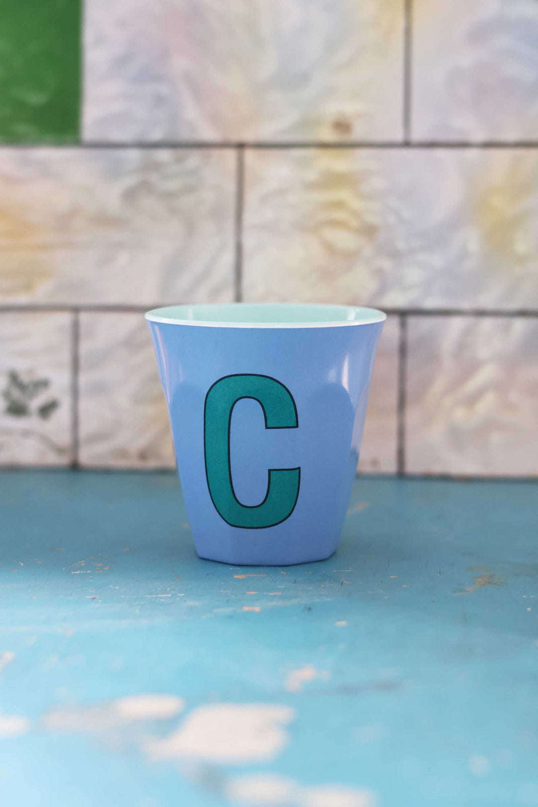 Blue cup with a green letter 'C' on a blue surface with a tiled wall background