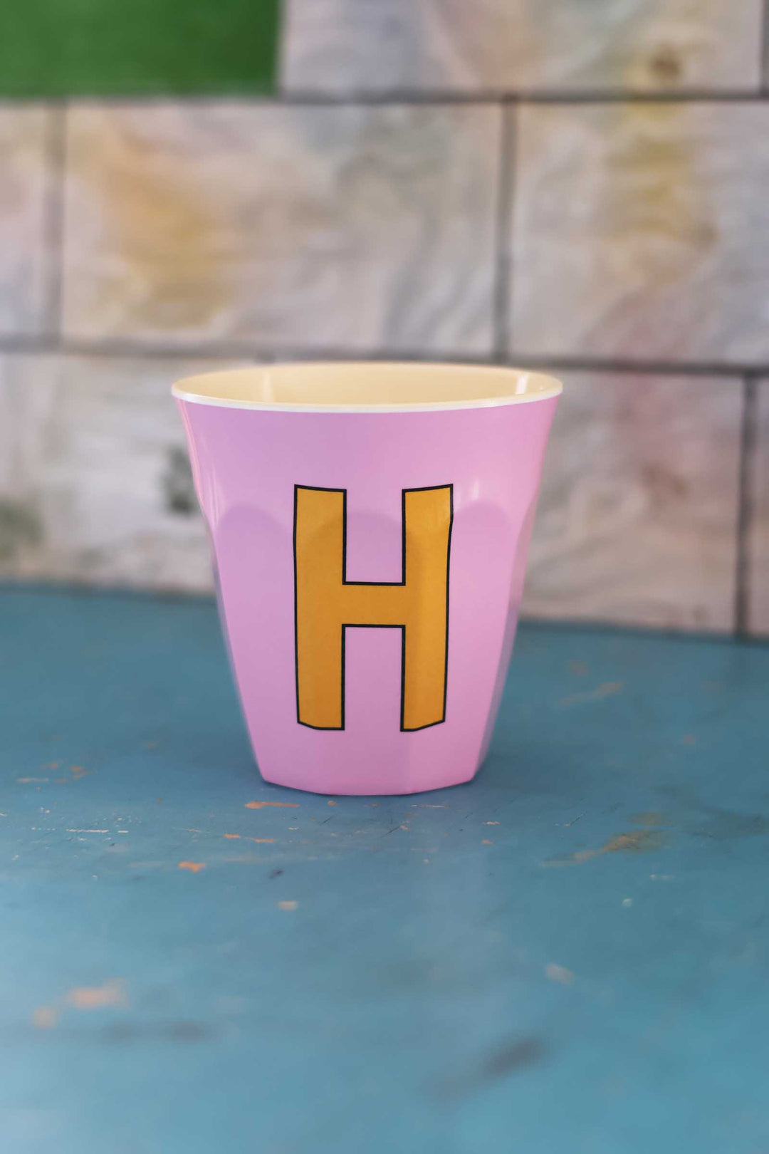 Pink cup with a yellow 'H' on a blue surface and stone wall background