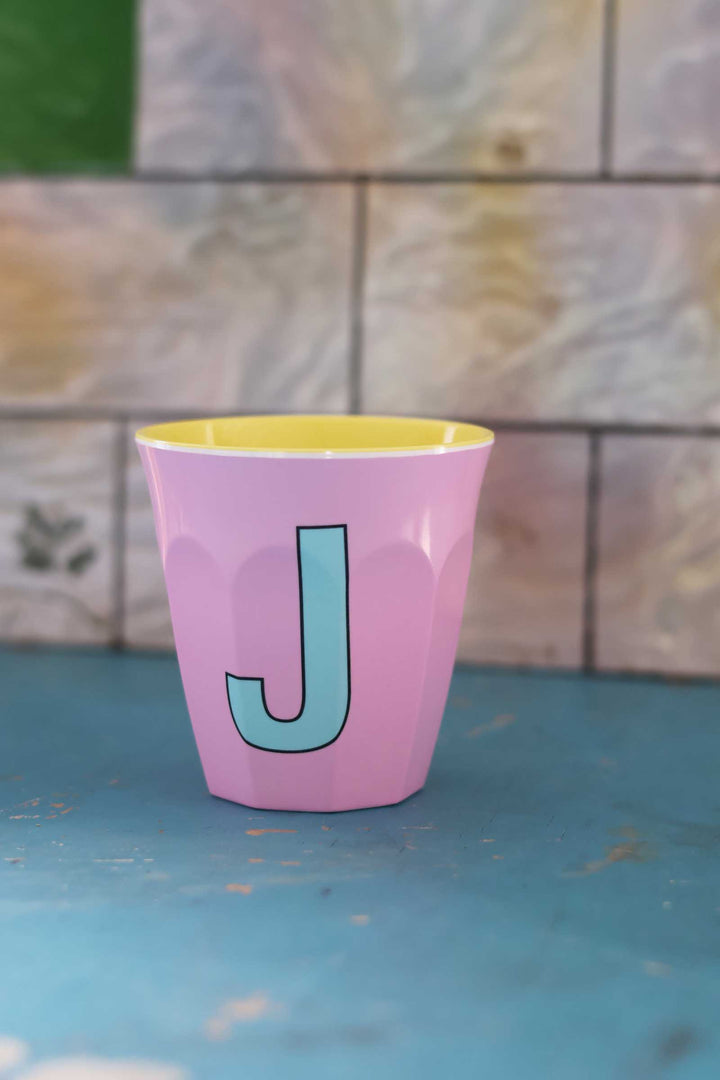 Pink cup with a yellow rim and blue letter 'J' on a blue surface with a tiled wall background.