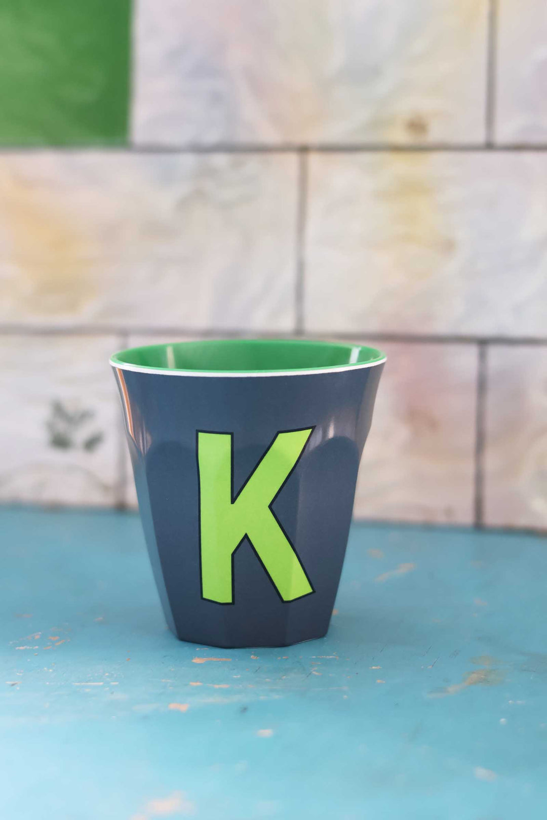 Green cup with a letter 'K' on a blue surface and tiled wall background