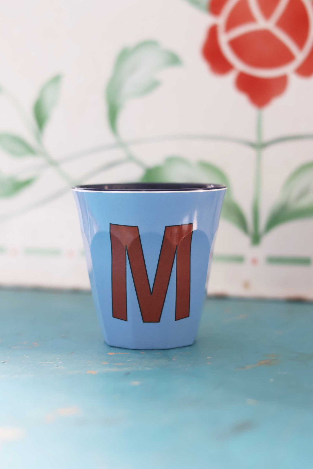Blue cup with brown letter 'M' on a light blue surface with floral background