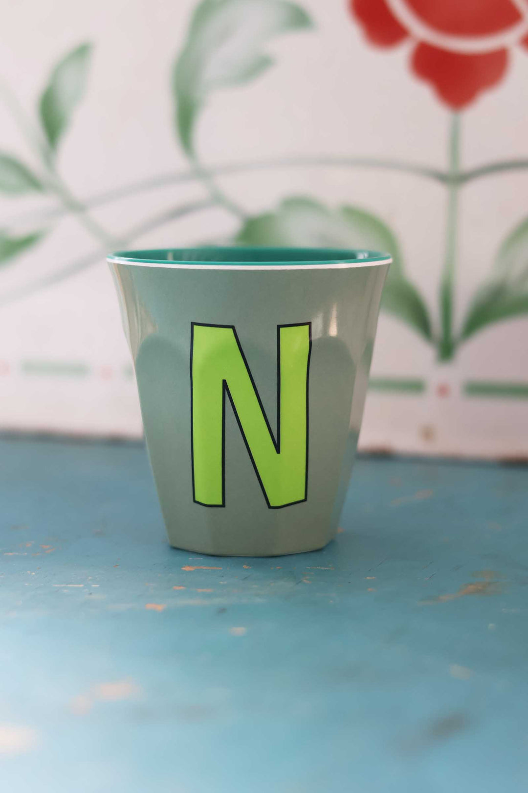 Ceramic cup with a green letter 'N' on a blue surface with a floral background