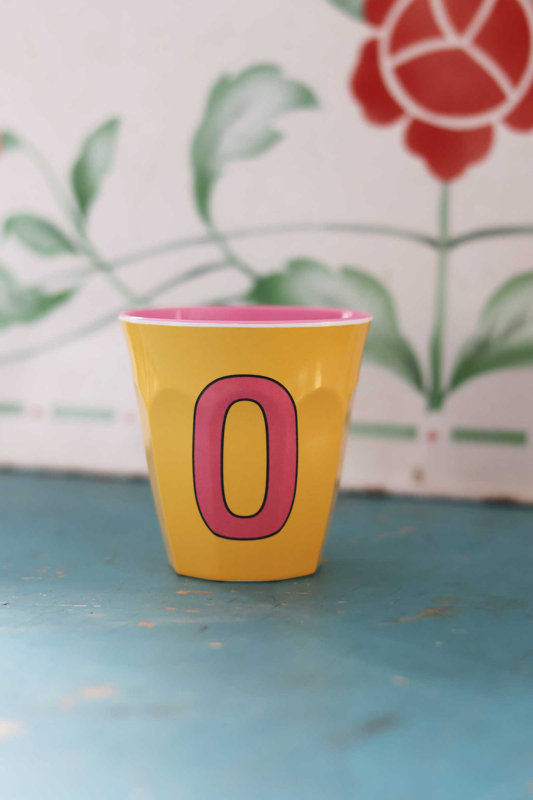 Yellow cup with pink interior and letter 'O' on a blue surface with floral background