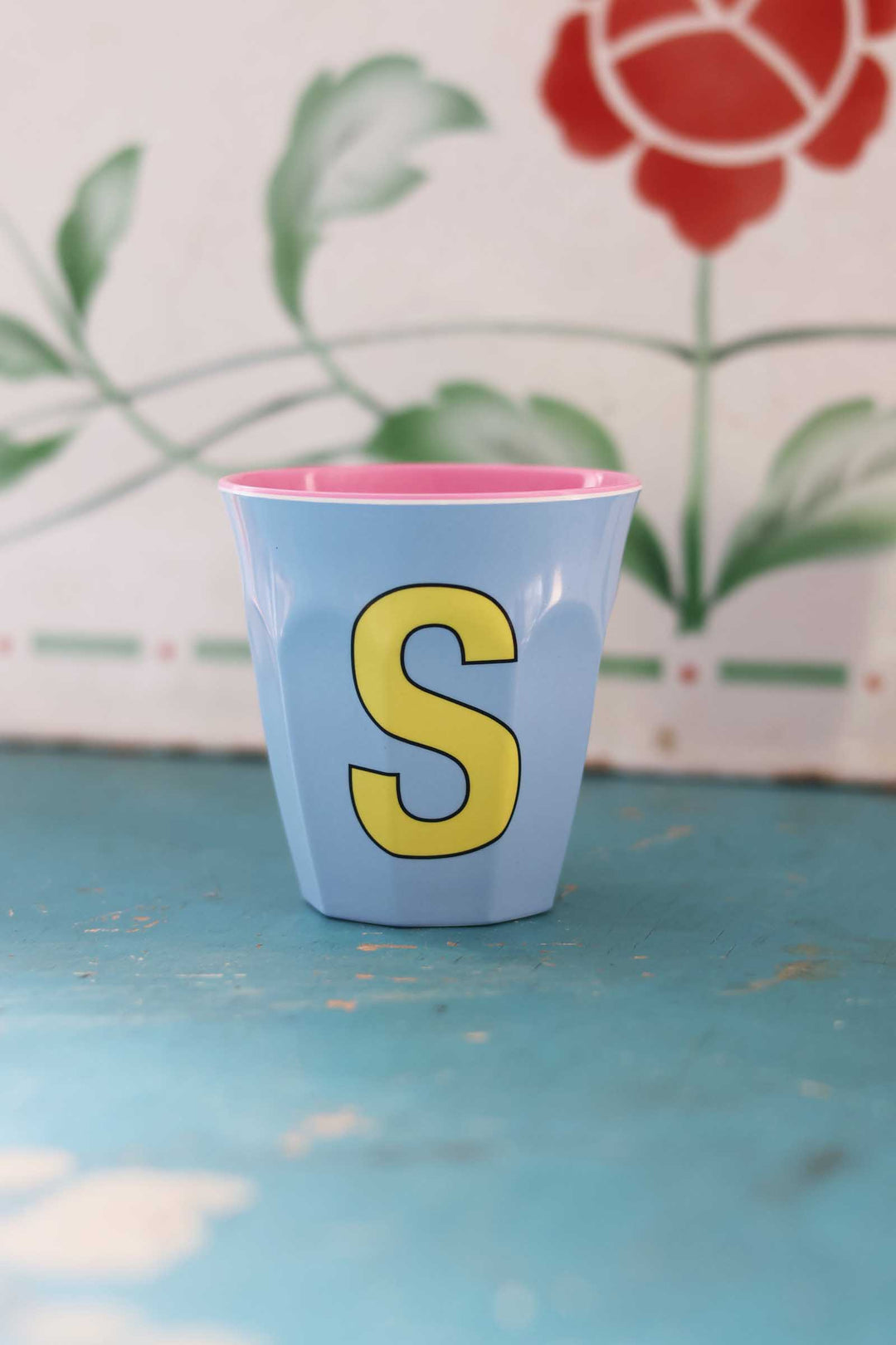 Blue cup with yellow letter 'S' on a floral-patterned background