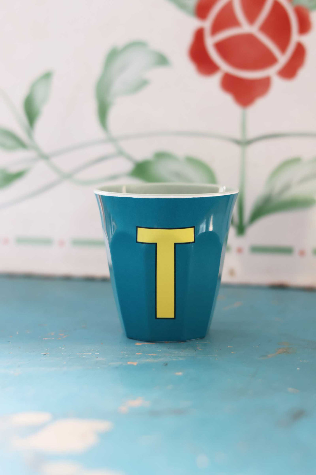 Blue cup with yellow letter 'T' on a light blue surface with floral background