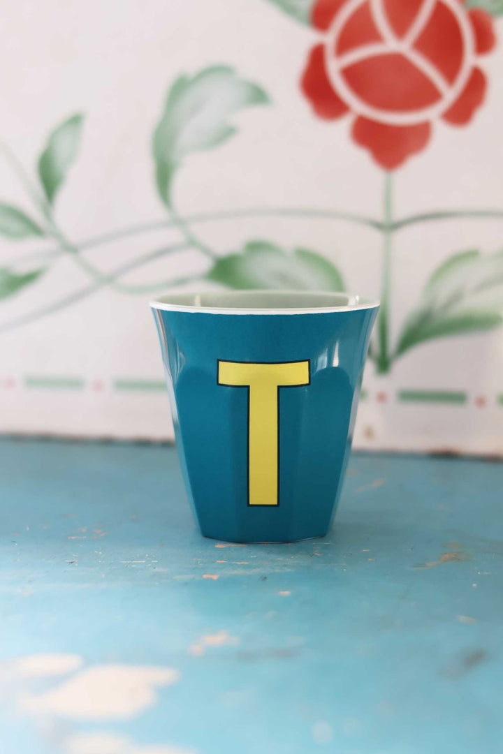 Blue cup with yellow letter 'T' on a light blue surface with floral background