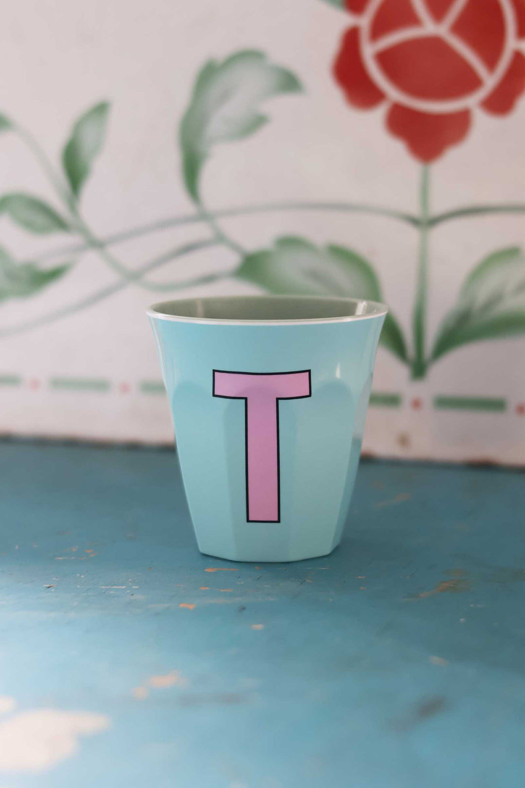Light blue cup with pink letter 'T' on a textured surface with floral background