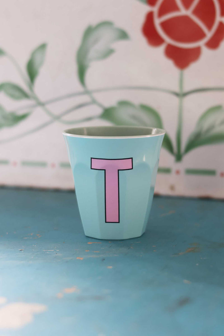 Light blue cup with pink letter 'T' on a textured surface with floral background