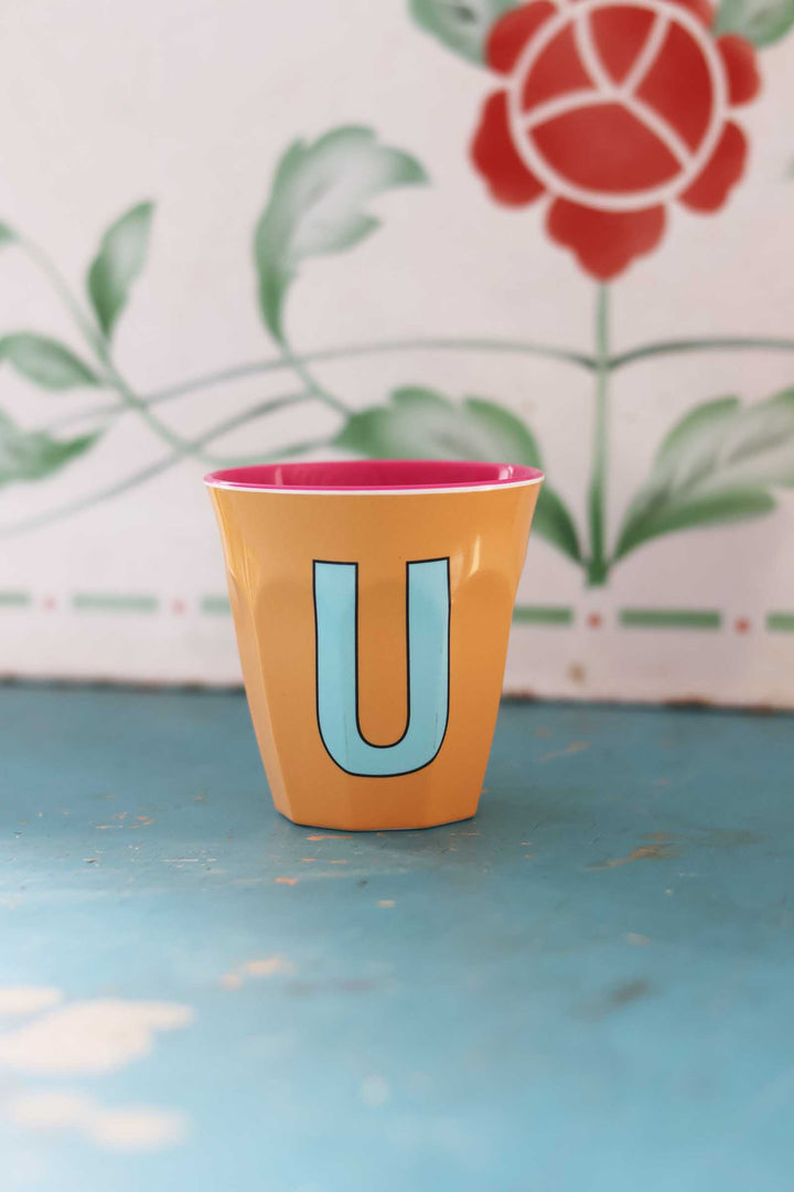 Yellow cup with a pink rim and blue letter 'U' on a blue surface with a floral patterned background.