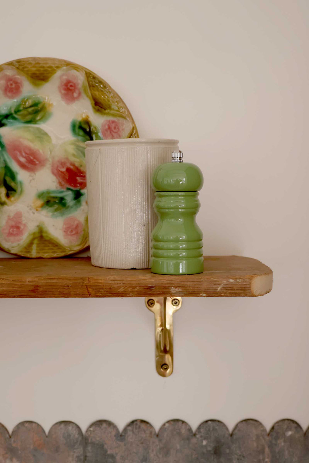 Green pepper grinder and white container on a wooden shelf with a decorative plate in the background.
