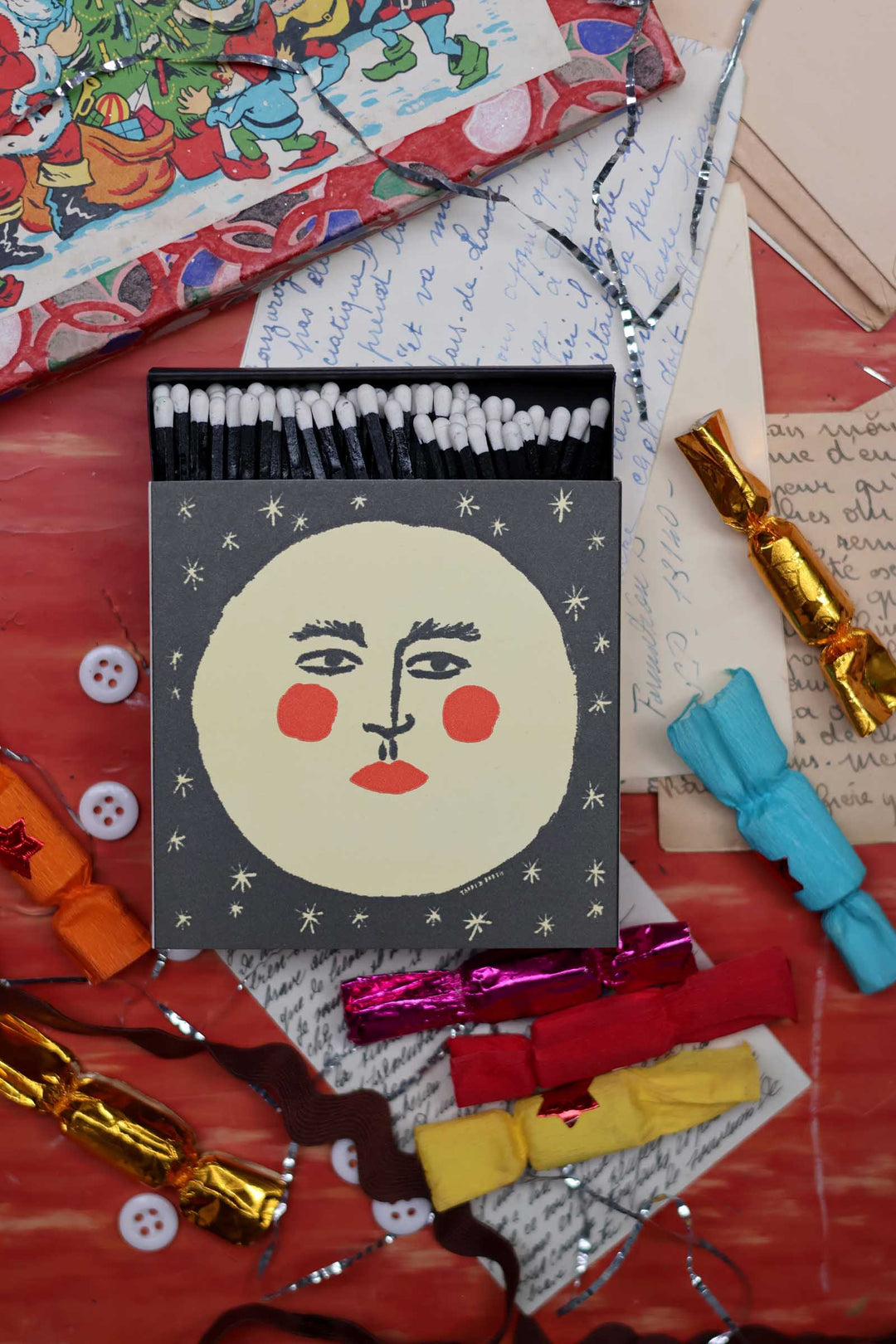 A square decorative matchbox with a stylized moon face design on the lid, surrounded by colorful cracker nuts, ribbons, buttons, and handwritten paper on a red tabletop.