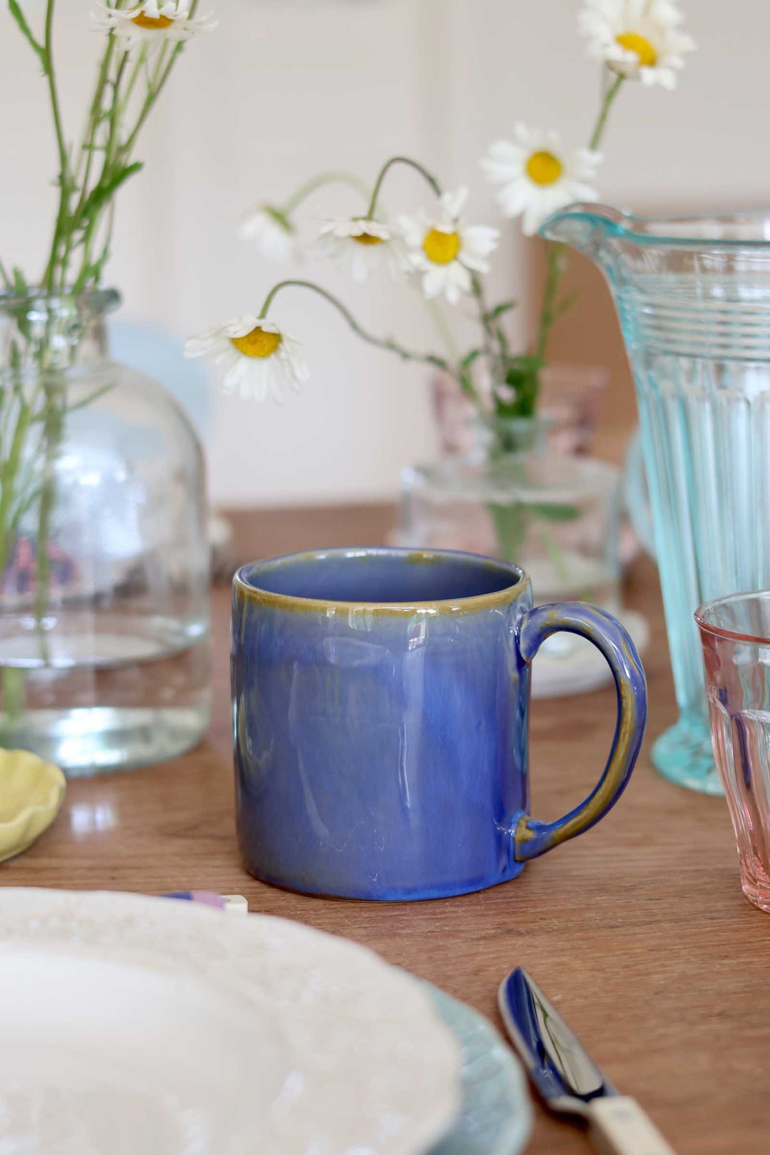 Blue mug on a wooden table with flowers and a glass in the background