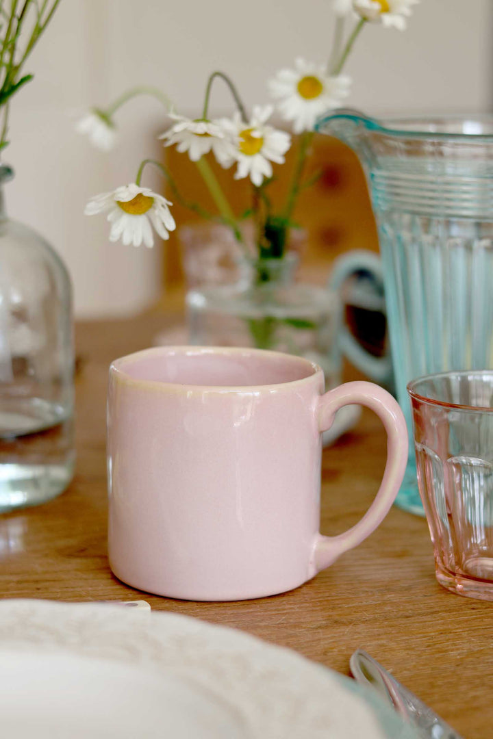 Pink mug on a wooden table with flowers and a glass pitcher in the background