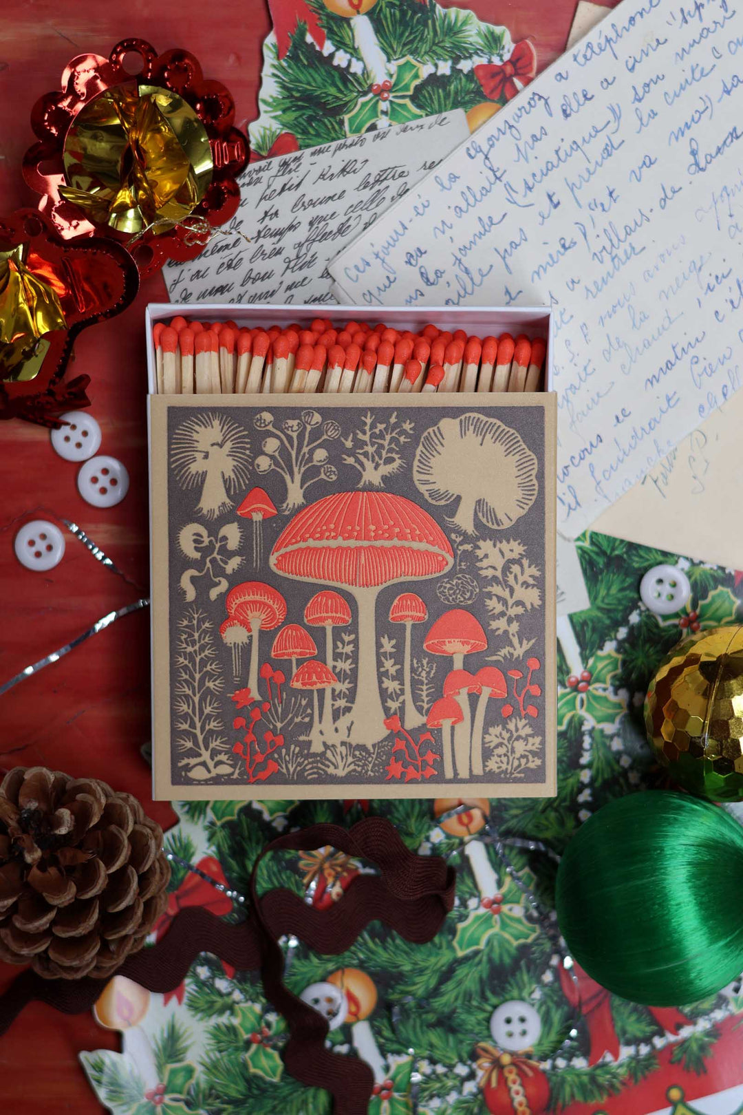 A vintage-style matchbox with illustrated red mushrooms on the lid, filled with red-tipped matches, arranged on a red holiday-themed surface with handwritten letters, ornaments, pinecones, and greenery.