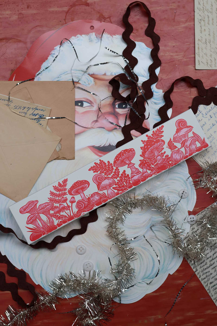 A styled flat-lay of Christmas-themed scrapbook paper featuring a vintage Santa Claus face, a red and white floral patterned strip, brown wavy ribbon, silver tinsel, and kraft paper envelopes on a textured red background.