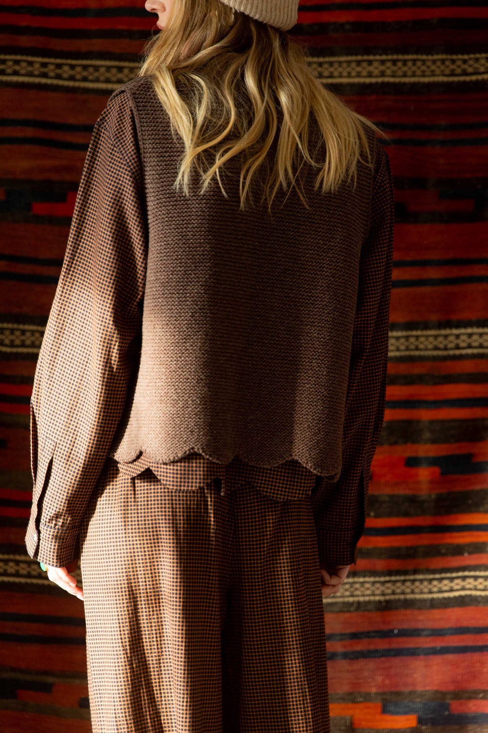 Person wearing a brown textured shirt and checkered pants standing against a colorful striped rug.