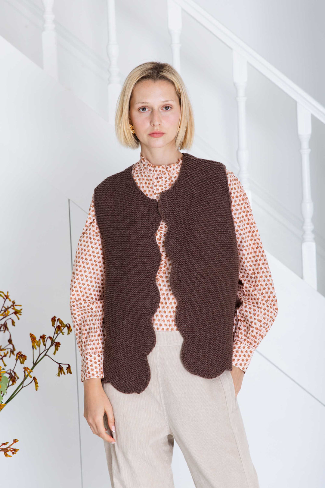 Woman wearing a brown knitted vest over a patterned shirt in a minimal indoor setting.