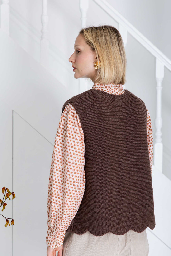 Woman wearing a brown knitted vest over a patterned shirt in a minimal indoor setting.