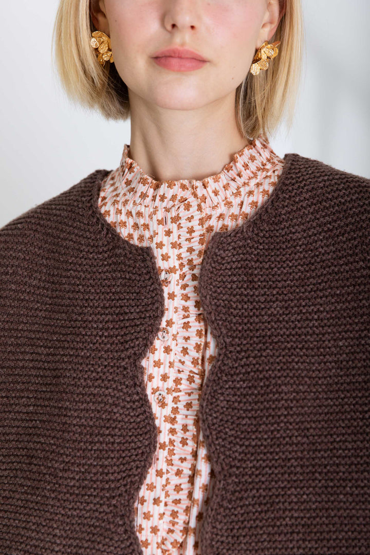 Close-up of a person wearing a brown knitted cardigan over a patterned blouse with a plain background.