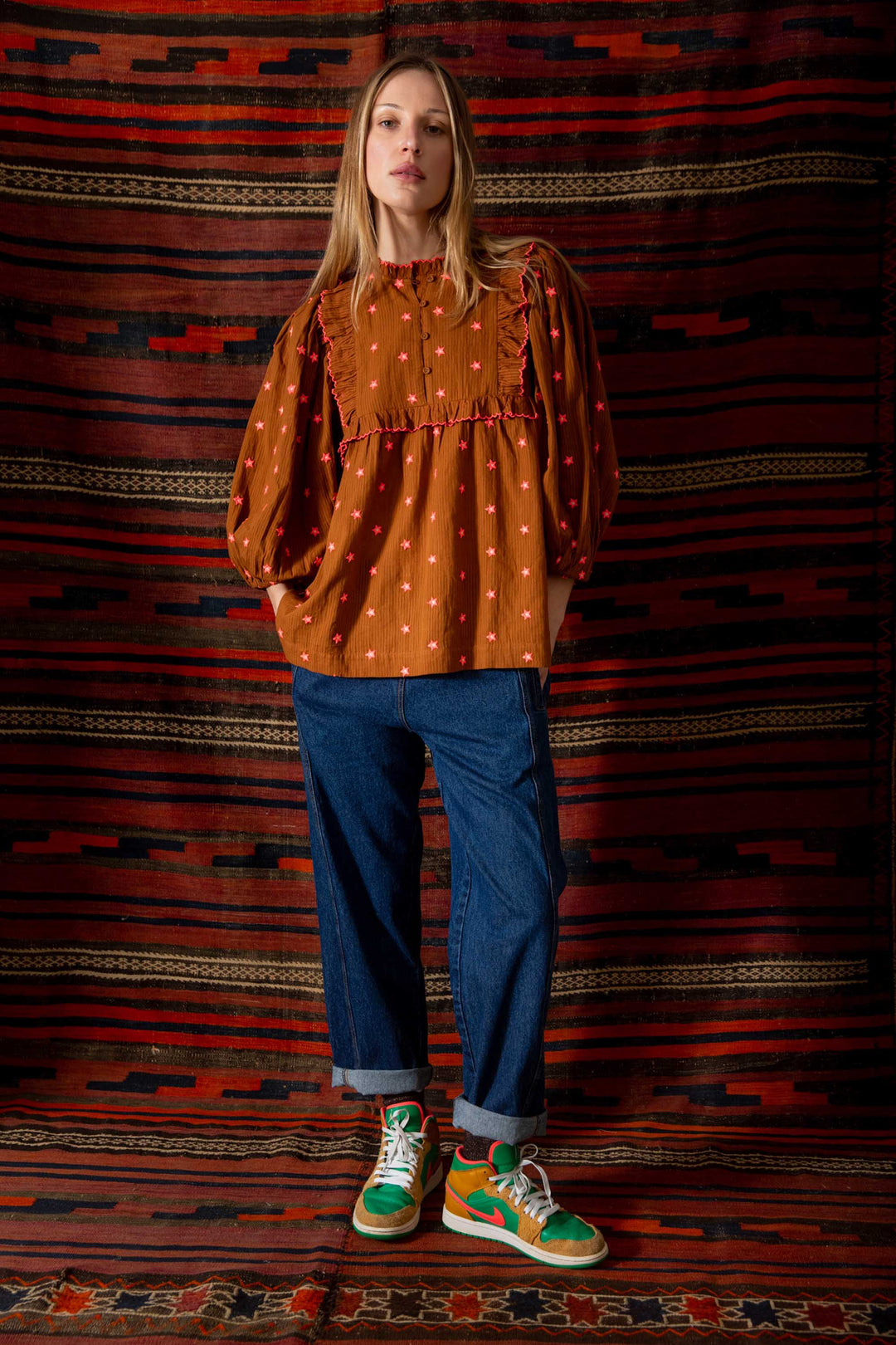 Person wearing a brown patterned top and blue jeans standing against a striped rug.