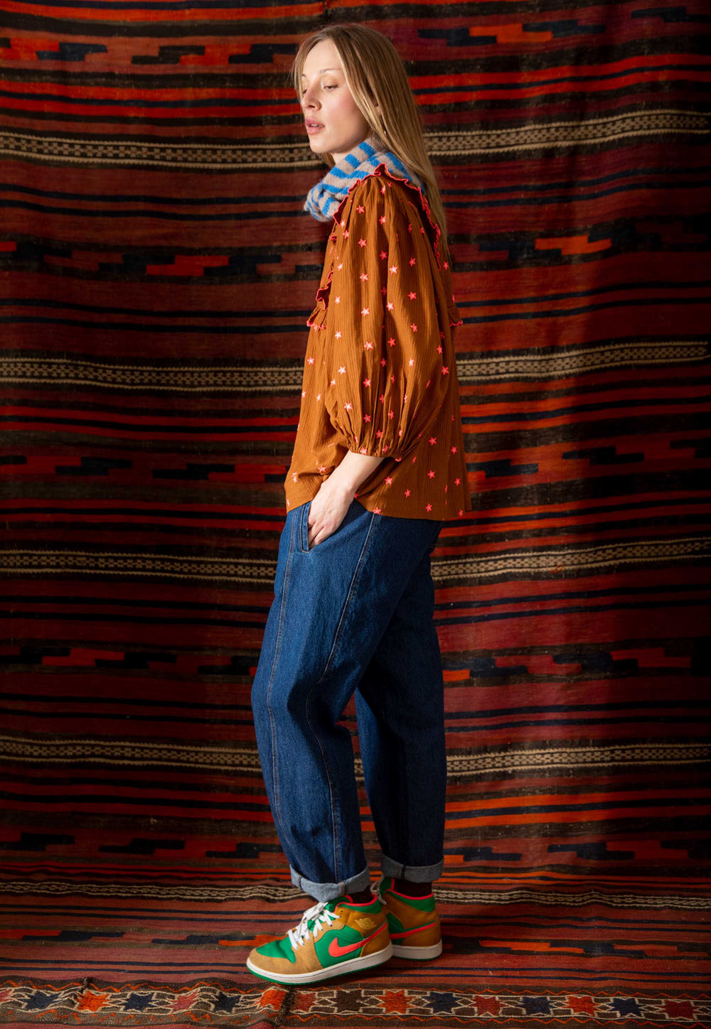 Person wearing a patterned shirt, jeans, and colorful sneakers against a textured wall.