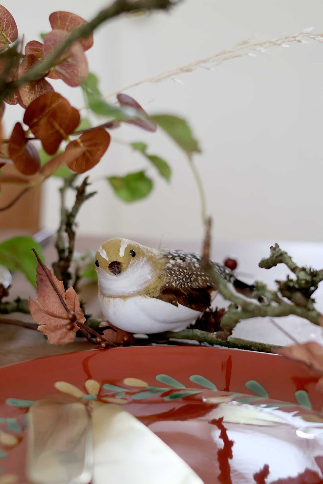 A small decorative bird figurine perched on a branch, styled with dried leaves and greenery in front of a red patterned plate.