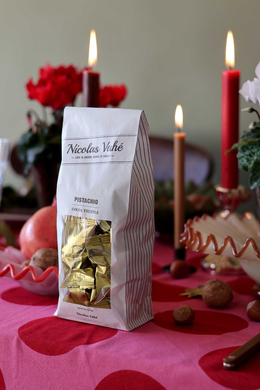 A bag of Nicolas Vahé Pistacchio Tacchino Treble chocolates stands on a pink patterned tablecloth surrounded by pomegranates, walnuts, and lit red candles in a warm, festive setting.