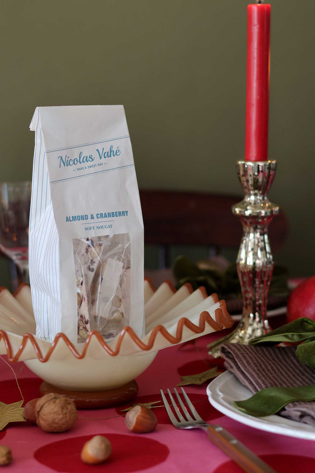A bag of Nicolas Vahé Almond & Cranberry Muffins sits on a decorated table setting with a red candle, plate, fork, walnuts, and green leaves.