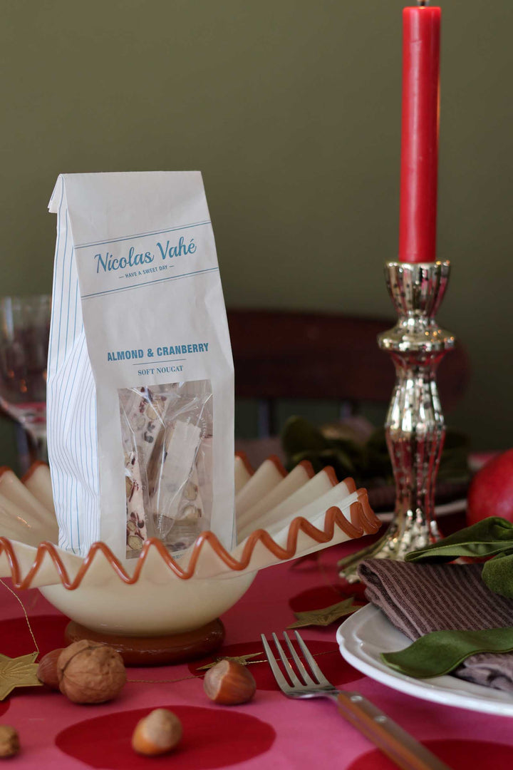 A bag of Nicolas Vahé Almond &amp; Cranberry Muffins sits on a decorated table setting with a red candle, plate, fork, walnuts, and green leaves.