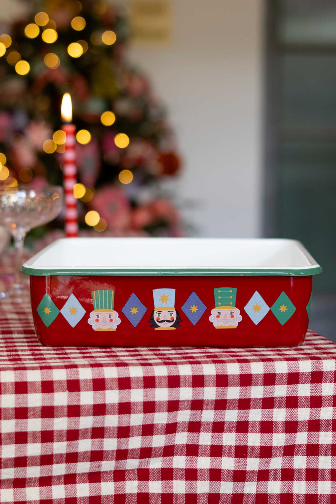 A red rectangular baking pan with a white interior and a green rim, decorated with colorful nutcracker and diamond motifs around the bottom edge, sitting on a red and white checkered tablecloth with a blurred Christmas tree in the background.