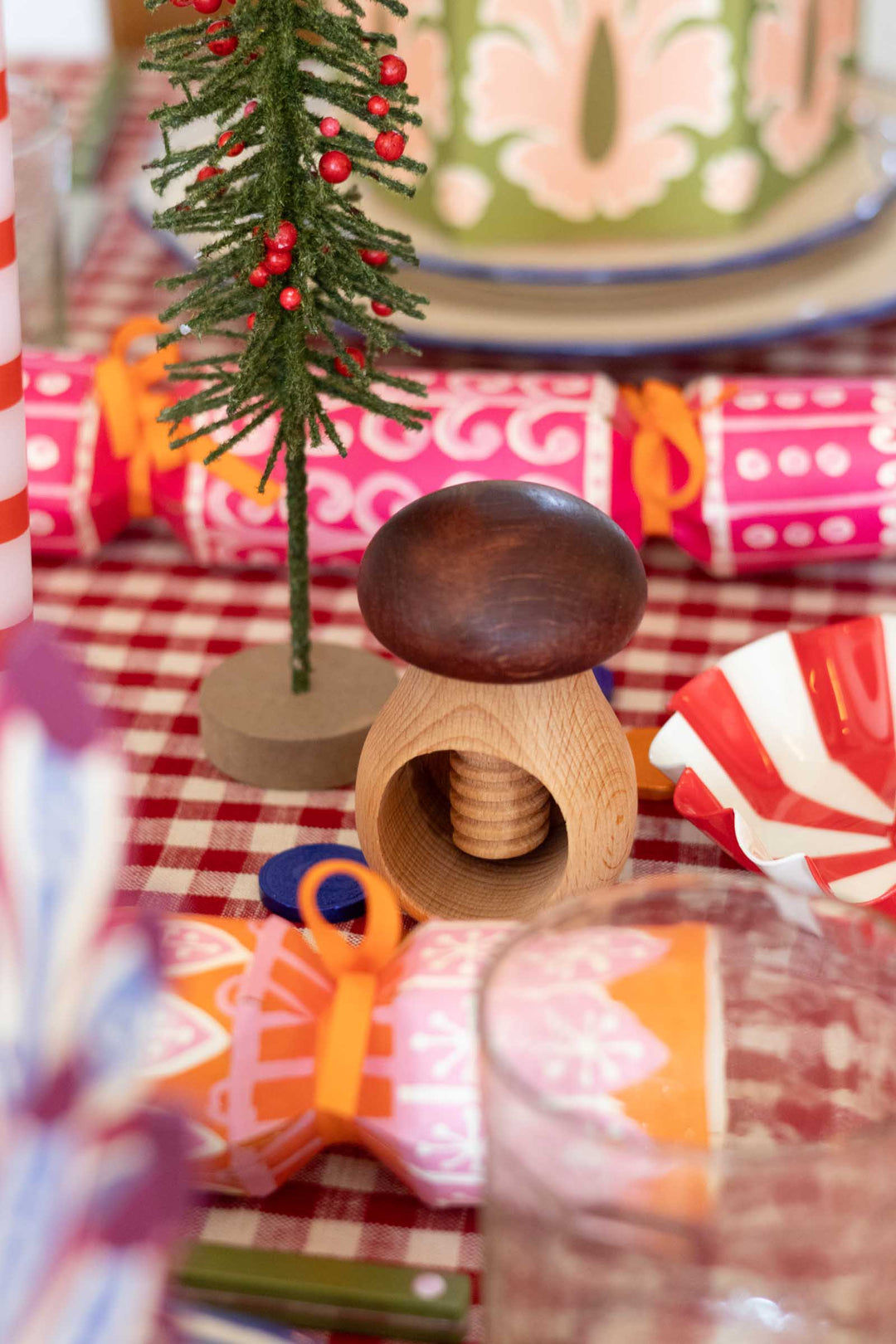 A festive tabletop scene with a small wooden mushroom-shaped ornament in the center, surrounded by Christmas decorations including a miniature evergreen tree with red berries, patterned cracker crackers, a red-and-white checkered tablecloth, and blurred holiday items in the background.