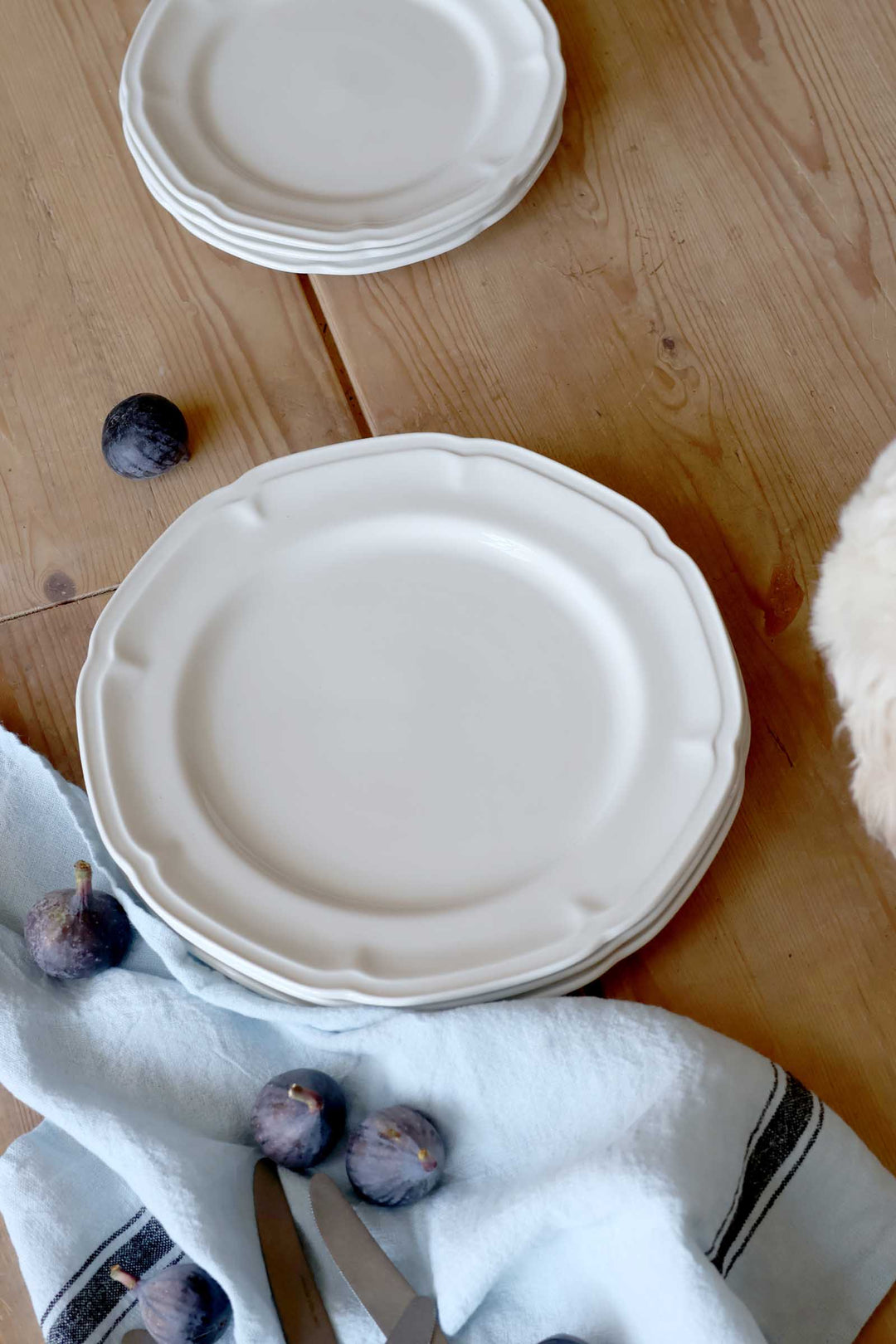 Stack of beige ceramic plates on a wooden surface with figs and a white towel.
