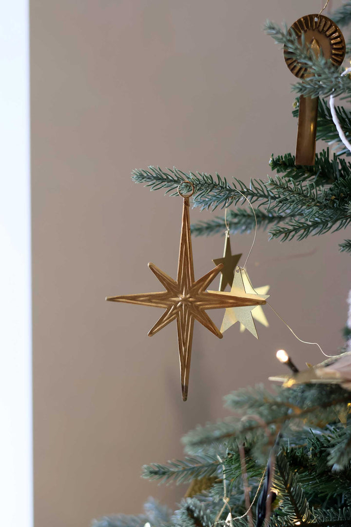 A gold metal star Christmas ornament hanging from a green Christmas tree branch, with warm string lights and other gold ornaments in the background.