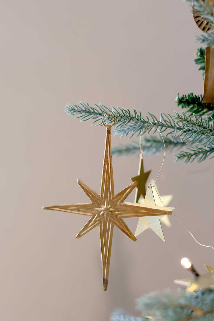 A gold metal star Christmas ornament hanging from a frosted green Christmas tree branch against a soft beige background.