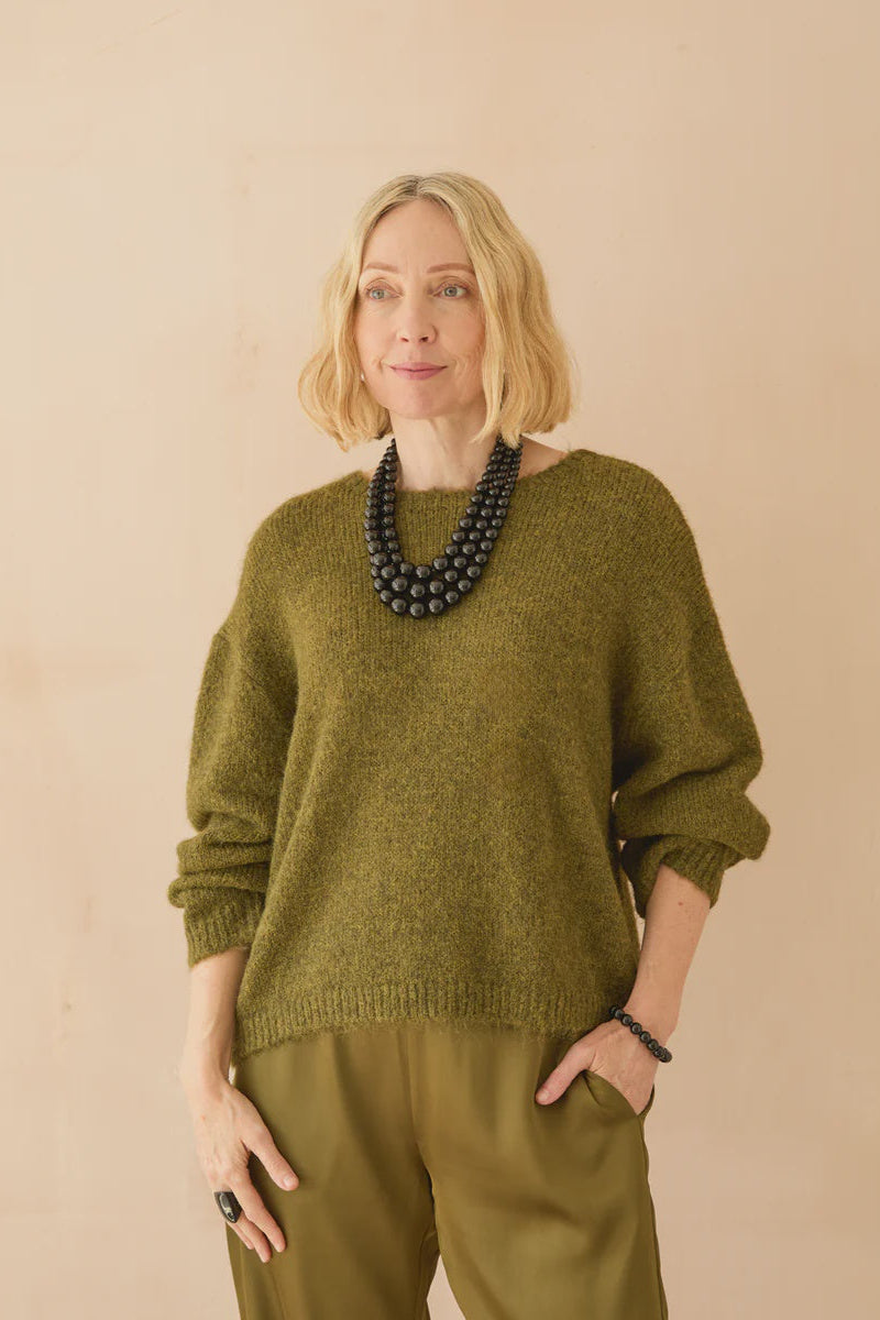 Woman wearing a green sweater and pants with a black necklace against a beige background