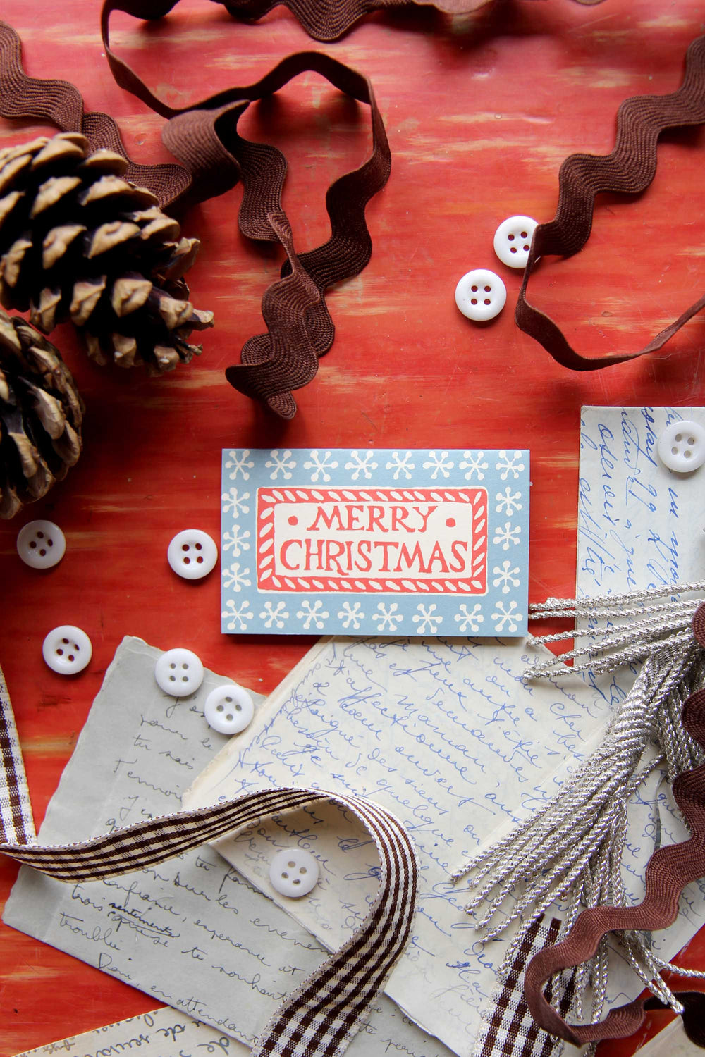 Merry Christmas card with ribbons, buttons, and pine cones on a red surface