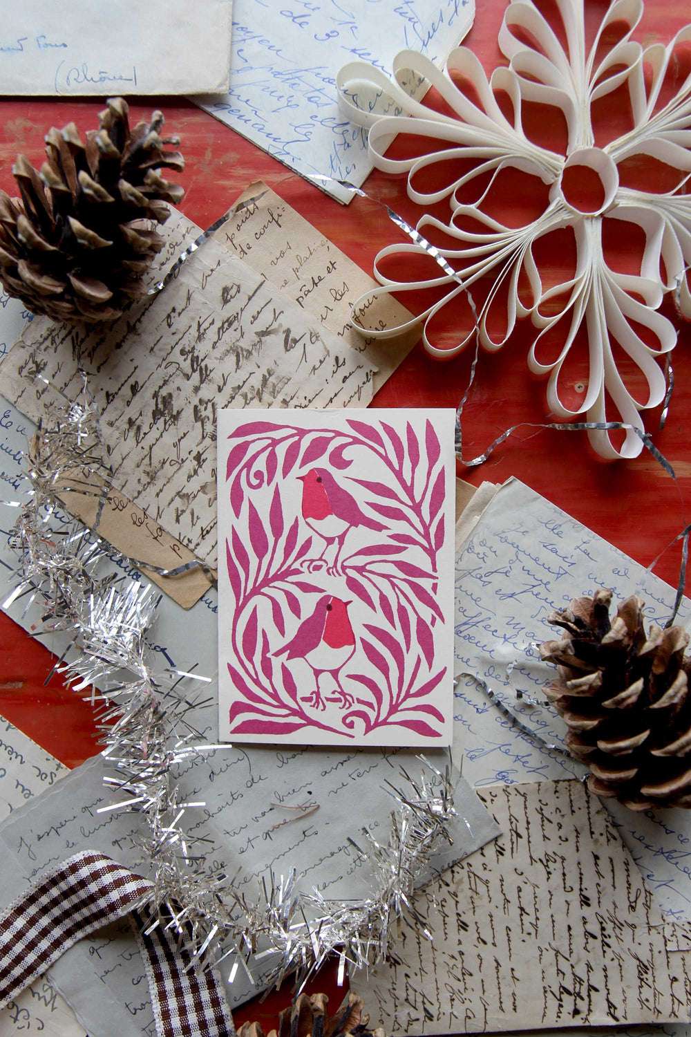 Decorative card with pink design on a red surface with pine cones and tinsel.