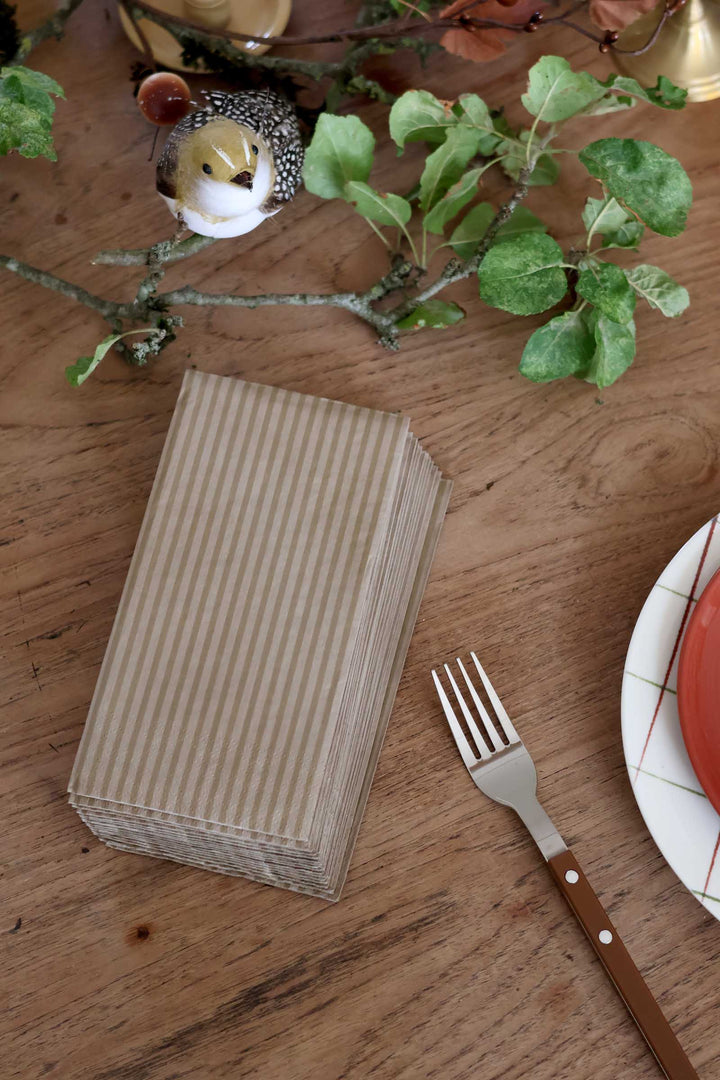 A stack of rectangular paper napkins with thin vertical beige and brown stripes sits on a wooden table, styled with a fork, a plate edge, green leaves, and a small decorative bird figurine.