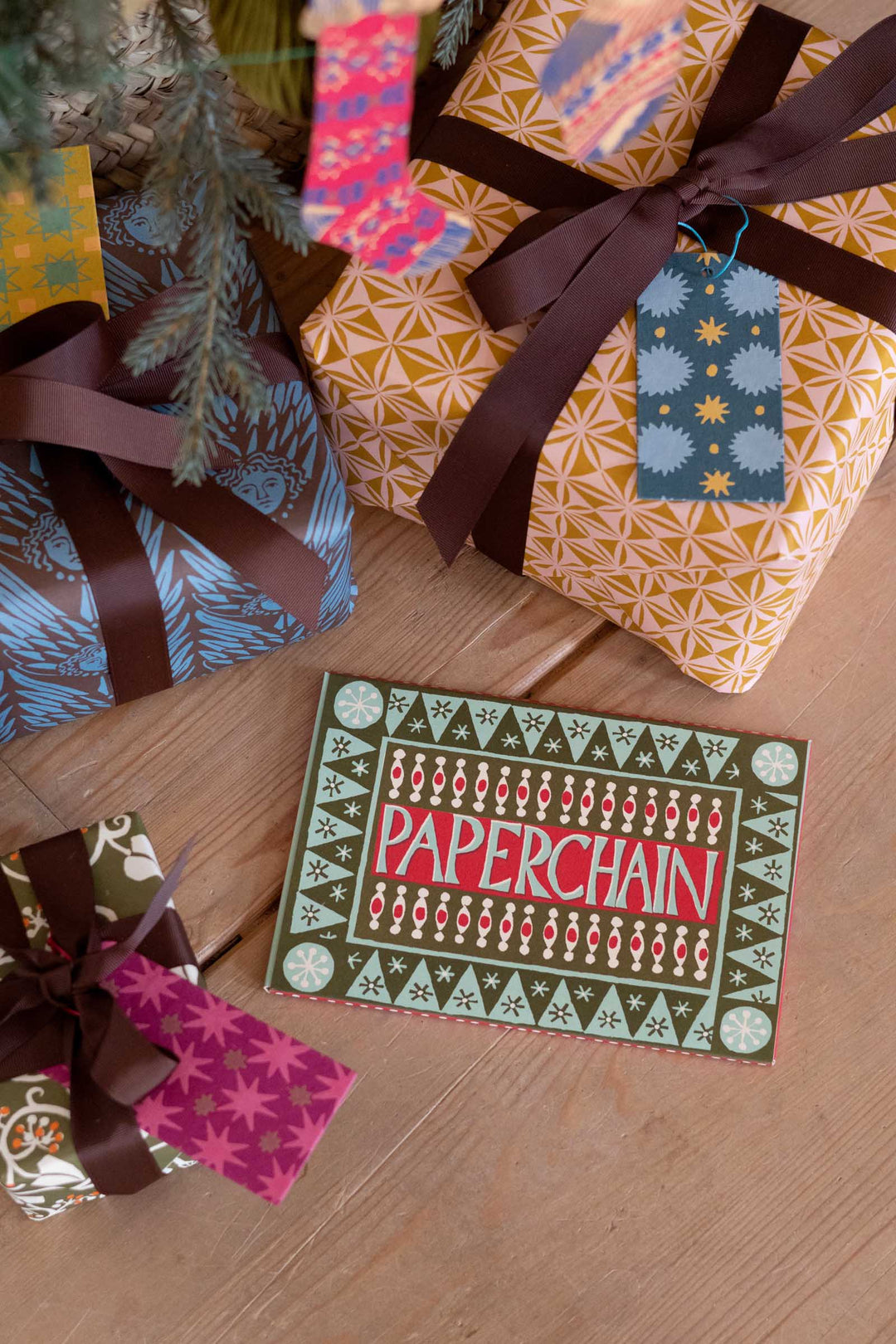 A Christmas-themed gift card with the word 'PAPERCHAIN' in red on a green patterned border, styled on a wooden floor among wrapped presents with brown ribbons and colorful wrapping paper.