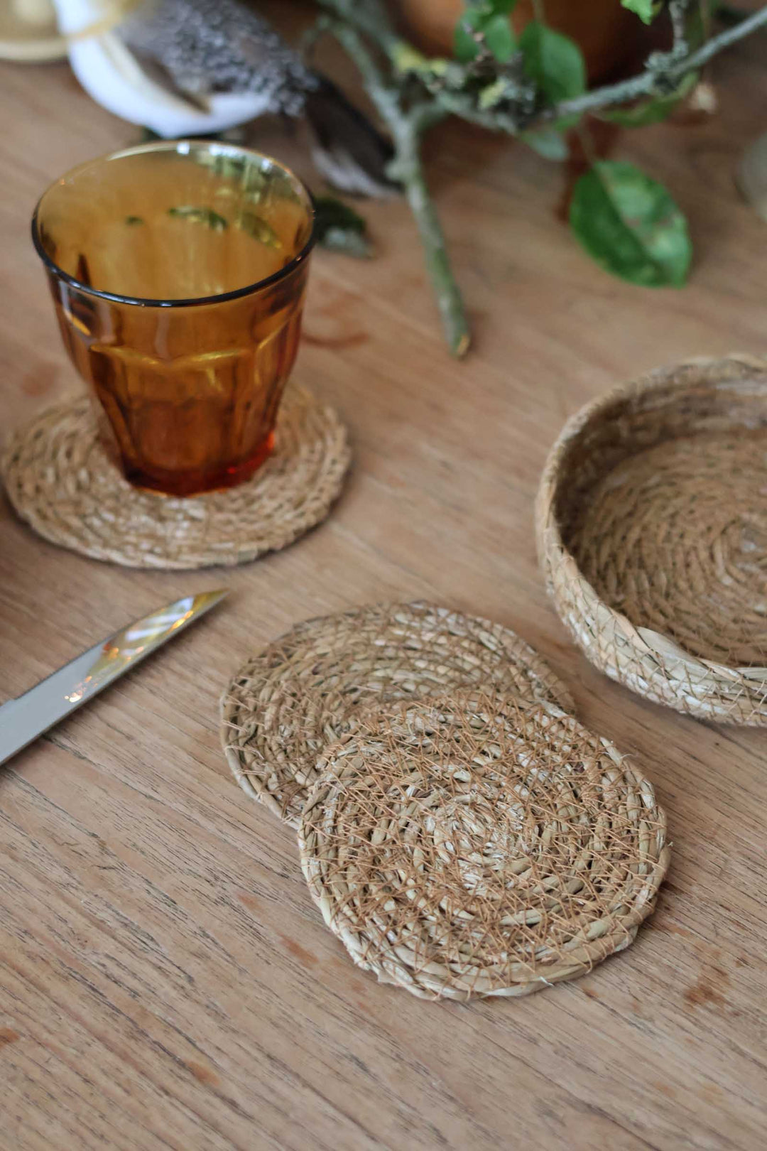Natural woven seagrass coasters and a matching small basket on a wooden table, with an amber glass tumbler resting on one coaster.