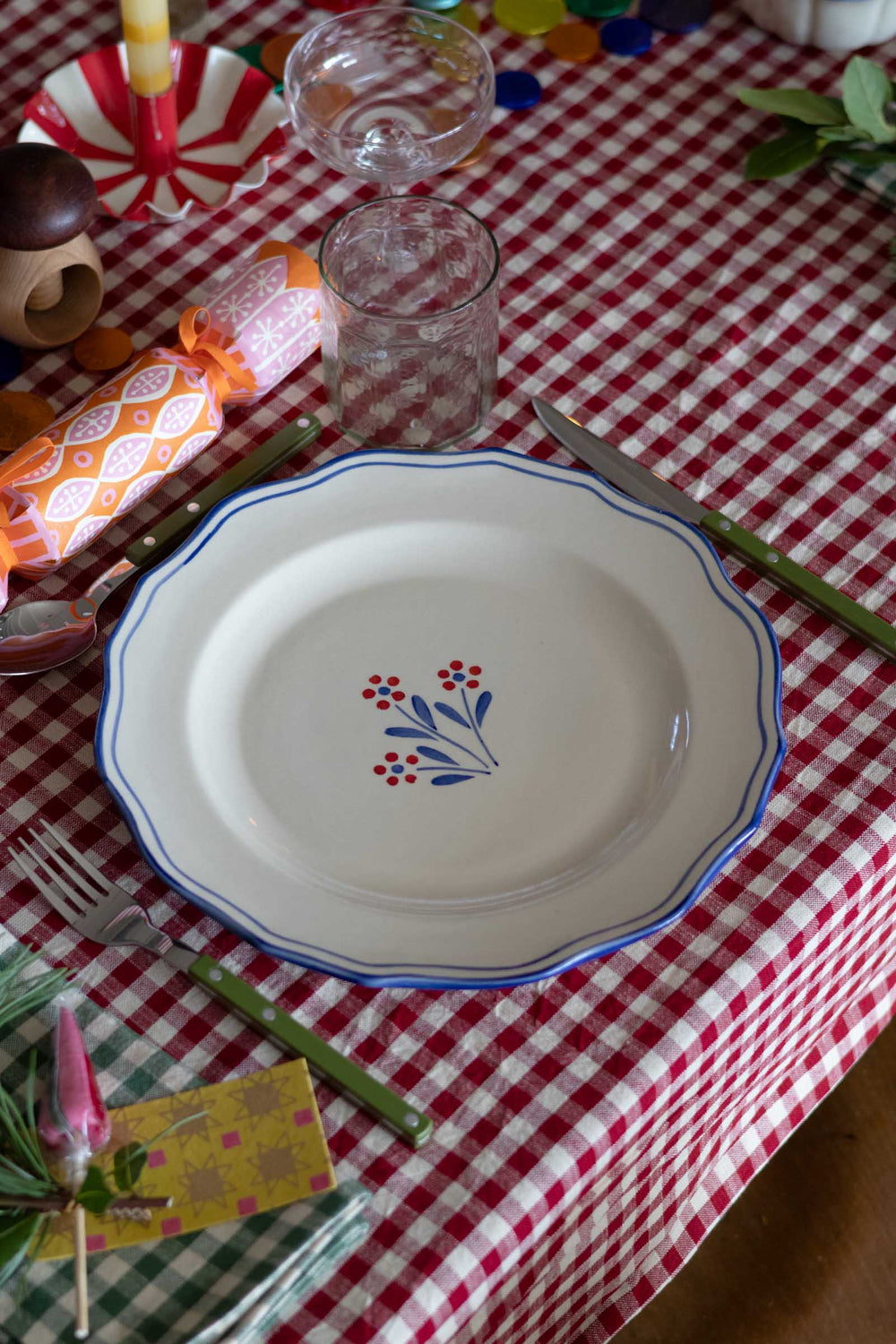 A white ceramic plate with a blue rim and a small floral motif in the center, set on a red and white checkered tablecloth with cutlery and other tableware.