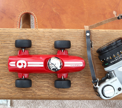 Red toy car with black wheels on a wooden surface next to a camera.