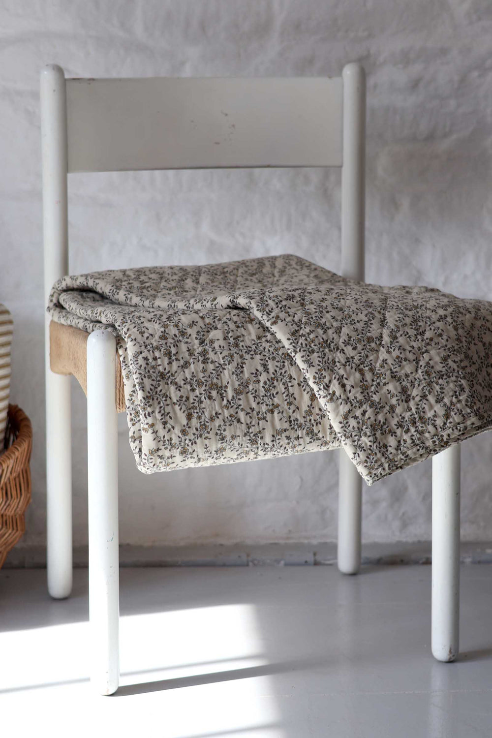 A textured, speckled beige and gray throw blanket is draped over a white wooden chair in a bright, minimalist room with a white brick wall.