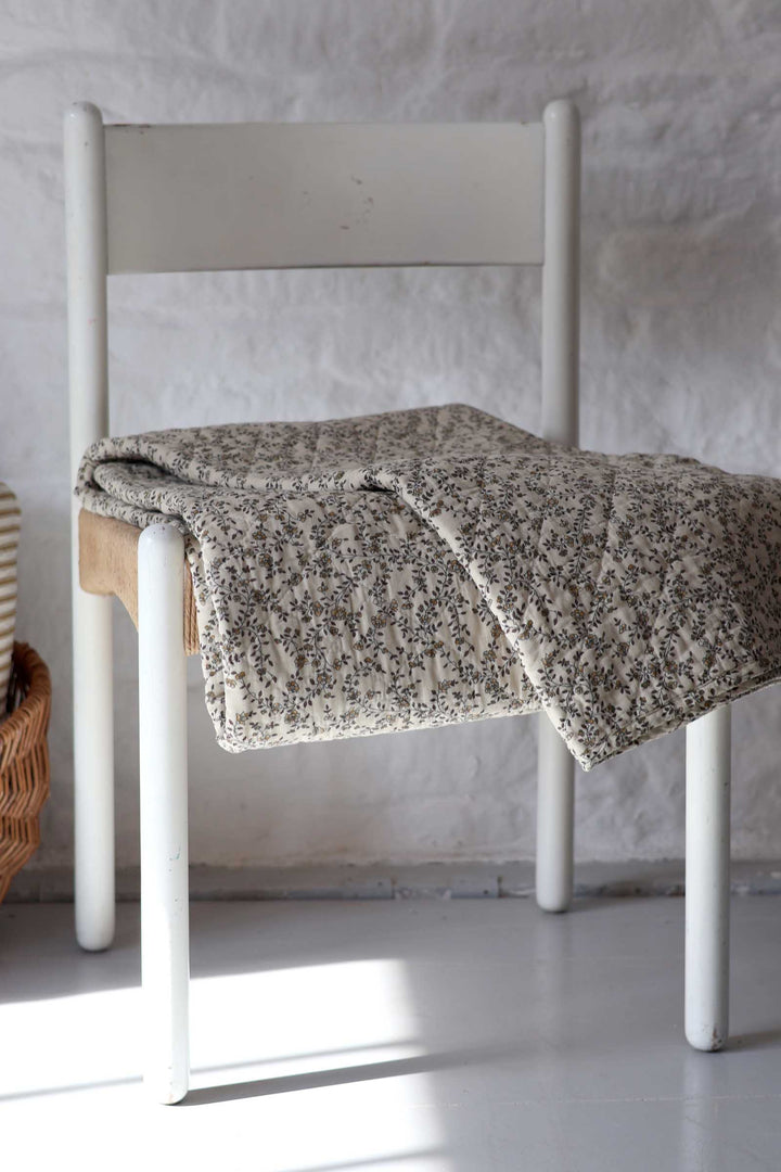 A textured, speckled beige and gray throw blanket is draped over a white wooden chair in a bright, minimalist room with a white brick wall.